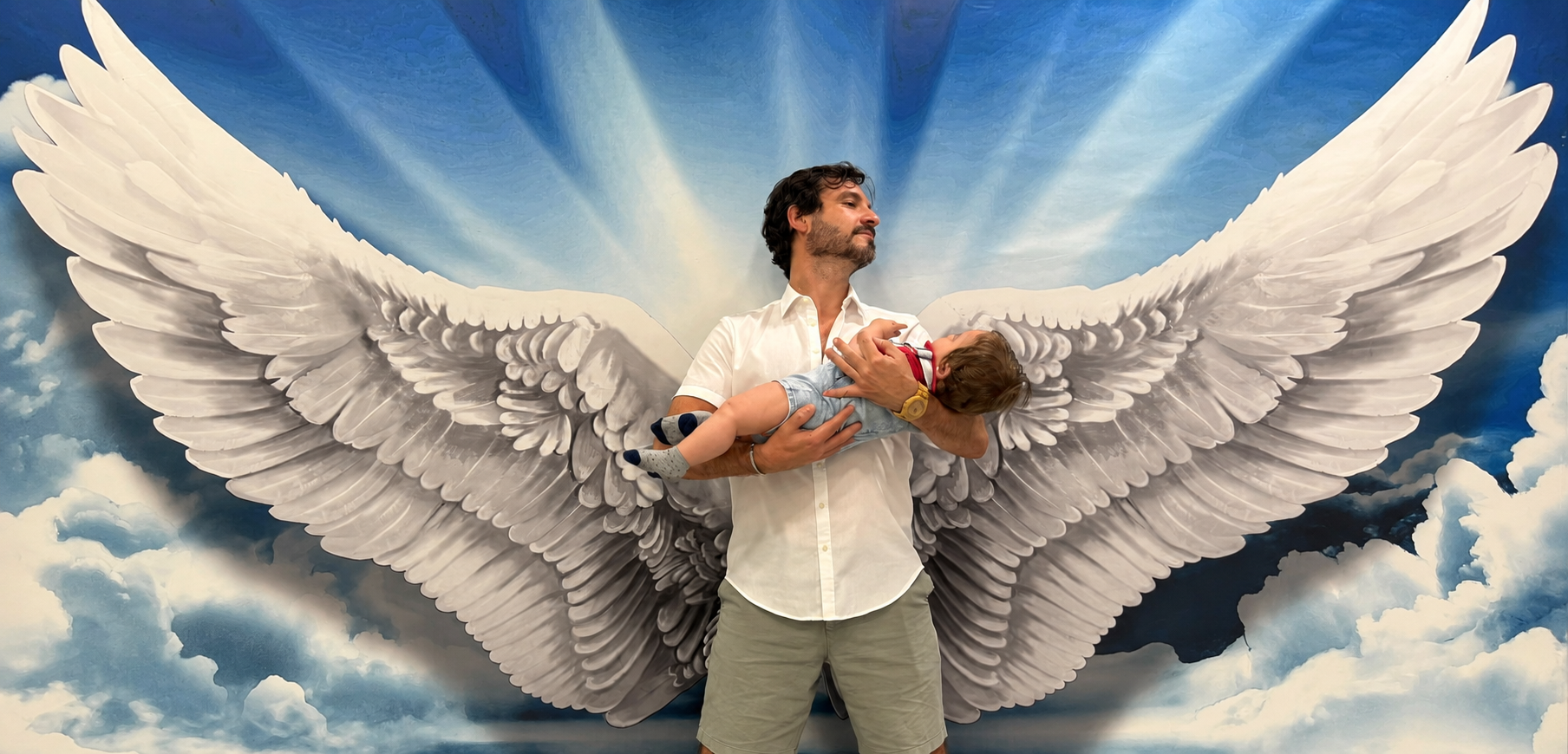 Alberto sosteniendo a un niño frente a un mural de grandes alas blancas